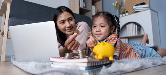 two kids sitting on the floor looking in front of a computer, looking at money with a piggy bank and coins next to them