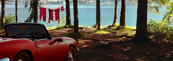 Red coverable car sitting under trees near a lake with Santa's clothing sitting hanging between the trees
